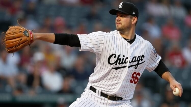 A baseball player in a white pinstripe uniform with "Chicago" on the chest and number 56 is pitching. He is mid-throw, holding a glove in one hand and an MLB ball in the other. The background shows a blurred crowd, capturing the energy that MLB maintains throughout the game.