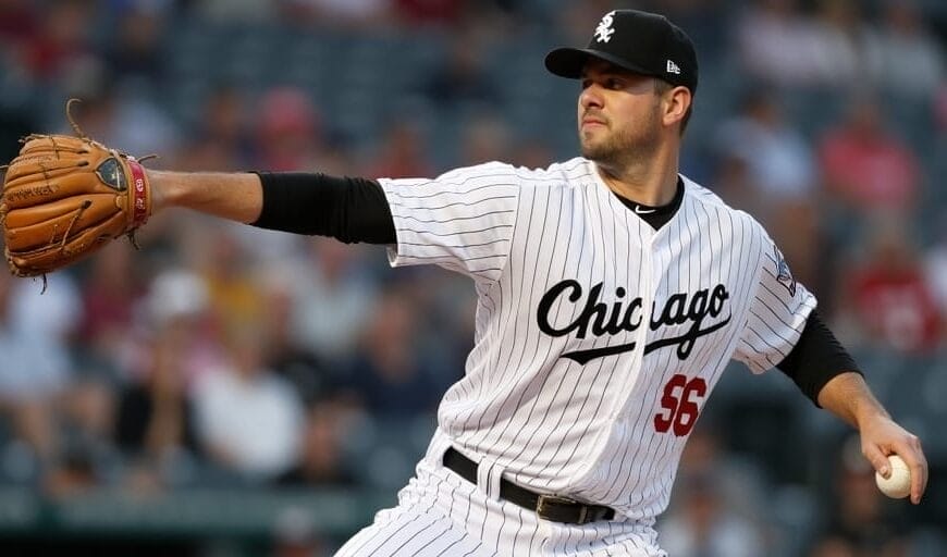 A baseball player in a white pinstripe uniform with "Chicago" on the chest and number 56 is pitching. He is mid-throw, holding a glove in one hand and an MLB ball in the other. The background shows a blurred crowd, capturing the energy that MLB maintains throughout the game.