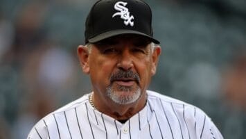 A man with a gray goatee wearing a black Chicago White Sox cap and a white pinstriped MLB jersey stands in a baseball stadium, with out-of-focus seats in the background—a nod to the league's two sets of rules.