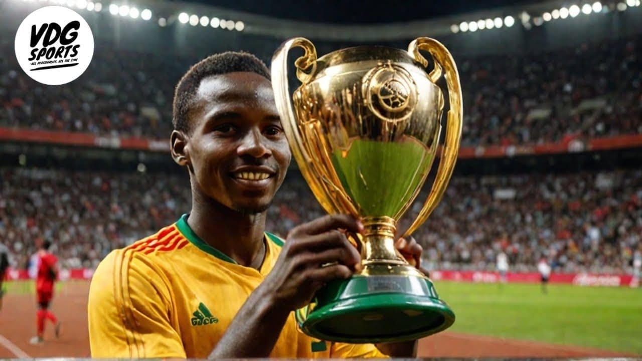 A man in a yellow sports jersey stands in a stadium holding a large golden trophy, celebrating his AFCON victory with the crowd in the background.