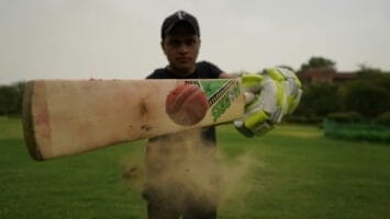 A person in a black cap and gloves hits a cricket ball with a bat on a grassy field, causing dust to fly, as fans cheer for this exciting moment of sports expansion.