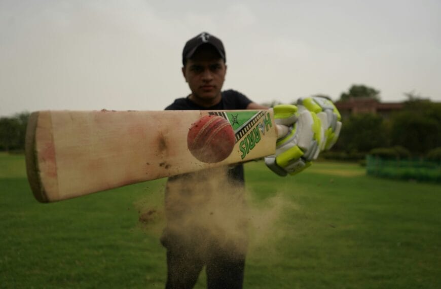 A person in a black cap and gloves hits a cricket ball with a bat on a grassy field, causing dust to fly, as fans cheer for this exciting moment of sports expansion.