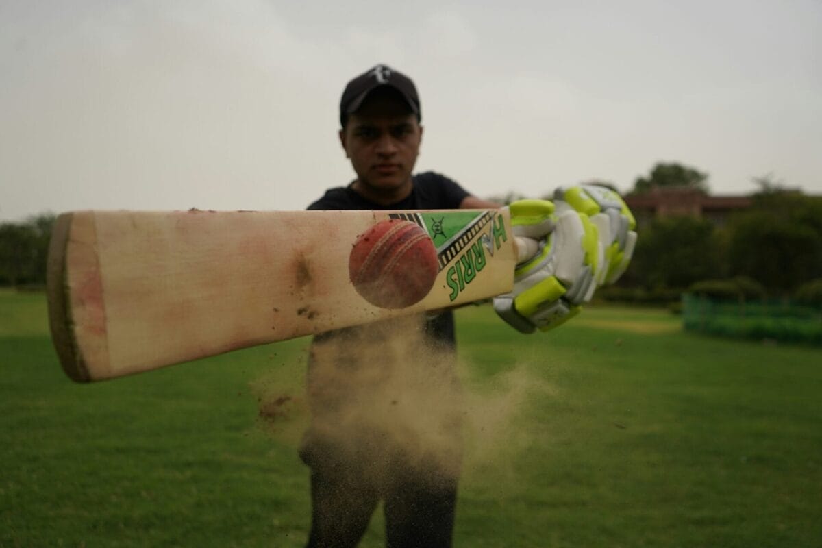 A person in a black cap and gloves hits a cricket ball with a bat on a grassy field, causing dust to fly, as fans cheer for this exciting moment of sports expansion.