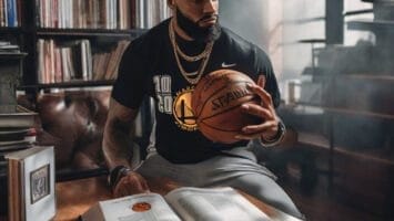 A bearded man in a sports jersey sits in a library, holding a basketball and looking at an open book about NBA games on the table. Shelves of books and a window are in the background.