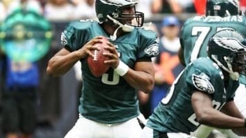 An NFL quarterback in a green uniform prepares to pass while a teammate blocks, all set against the bustling crowd in the background.