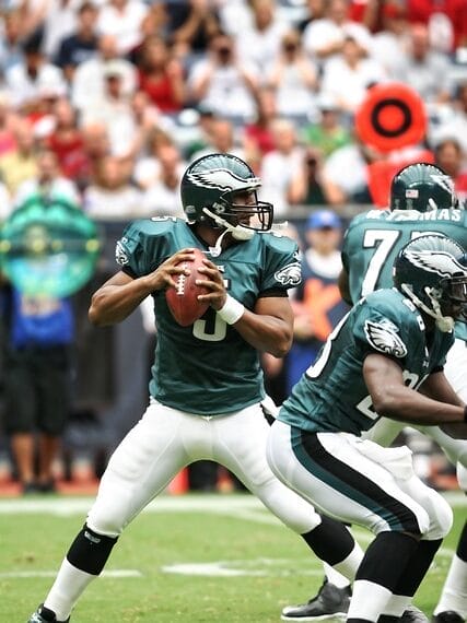 An NFL quarterback in a green uniform prepares to pass while a teammate blocks, all set against the bustling crowd in the background.