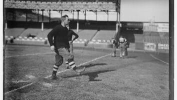 A vintage photo captures an early NFL quarterback in classic football attire, standing on a field with a grand stadium in the background.