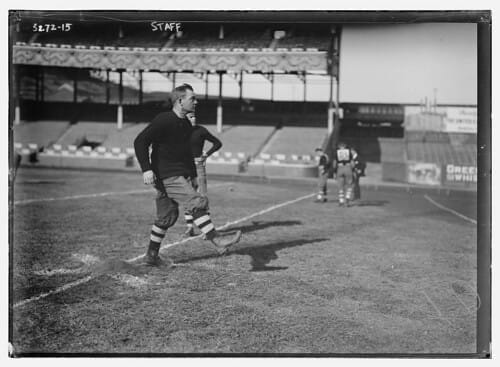 A vintage photo captures an early NFL quarterback in classic football attire, standing on a field with a grand stadium in the background.