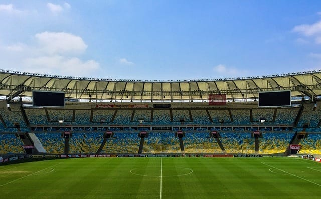 An empty stadium of soccer showcases a pristine green field, surrounded by tiered seating under a clear blue sky.