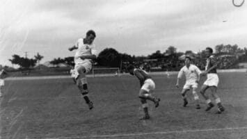 A vintage soccer match unfolds as a player skillfully jumps to head the ball, demonstrating the science behind soccer, while others watch with anticipation on an outdoor field.