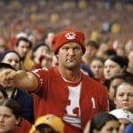 A focused man in a red hat and jersey, demonstrating emotional intelligence in sports fans by confidently pointing forward, surrounded by a crowd at a sports event.