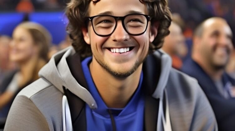 A young man with curly hair and glasses smiling at a sports event, wearing a blue shirt and grey jacket claims to know the smartest sports fans.
