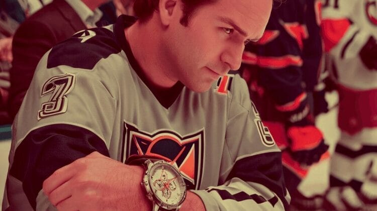 A man in a hockey jersey sits on a bench, resting his arms on his knees, with a serious expression. Players and spectators are visible in the background.