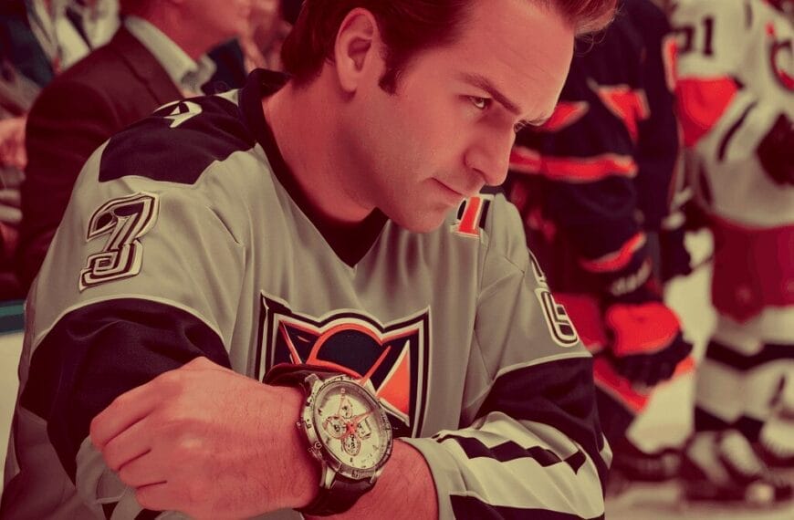 A man in a hockey jersey sits on a bench, resting his arms on his knees, with a serious expression. Players and spectators are visible in the background.