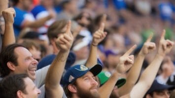 Crowd of sports fans in a stadium, wearing team jerseys and hats, cheer enthusiastically with their arms raised, creating an electrifying Battle of the Fans atmosphere.