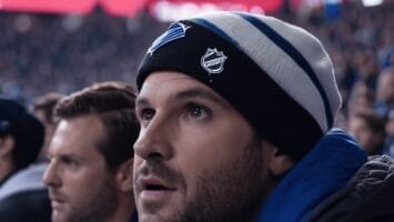 A man wearing a beanie with a hockey team logo watches the game intently from the stands, his focus unwavering. In the background, another man is equally engrossed in the action, making it clear that for these fans, MLS vs NHL is not even a question.