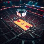 The empty indoor basketball arena echoes the legacy of NBA super teams, with a central court surrounded by rows of red seats and a scoreboard presiding above, ready for the next great match-up.