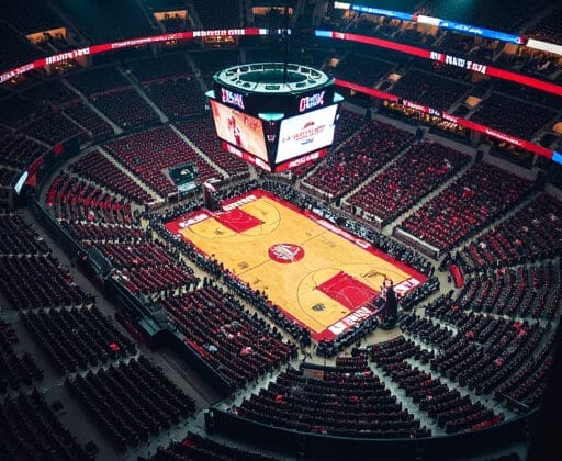 An empty basketball arena with red seats and a lit court awaits the thrill of NBA superteams. Scoreboards and banners hang proudly above the court, anticipating the next big game.