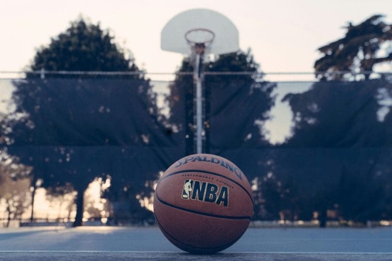A basketball with an NBA logo rests on an outdoor court, poised for the perfect dunk, with a hoop and trees gracefully framing the background.