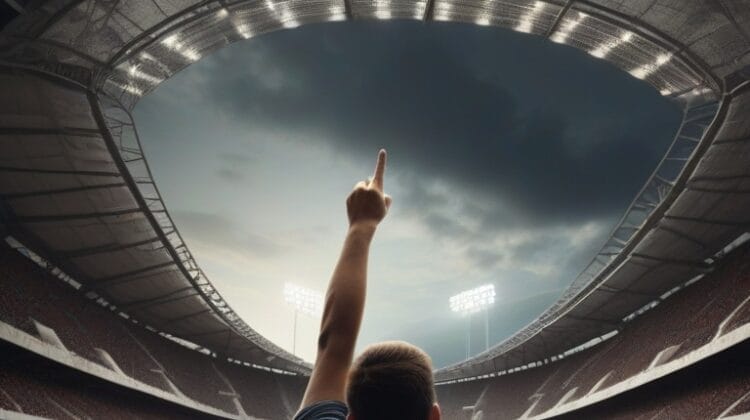 A person stands on a soccer field pointing upward towards the cloudy sky inside a large, illuminated stadium.