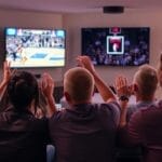 People watching an NBA game on TV, cheering with raised hands in a cozy living room, savoring the excitement of the sport purely for its thrills without analytics.