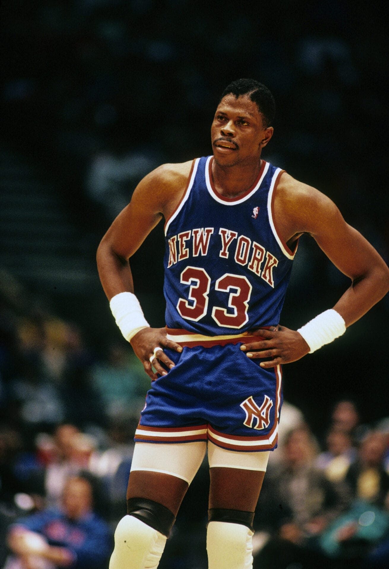 Basketball legend Patrick Ewing, donning his iconic blue New York jersey with the number 33, stands confidently with hands on hips during the intense game.