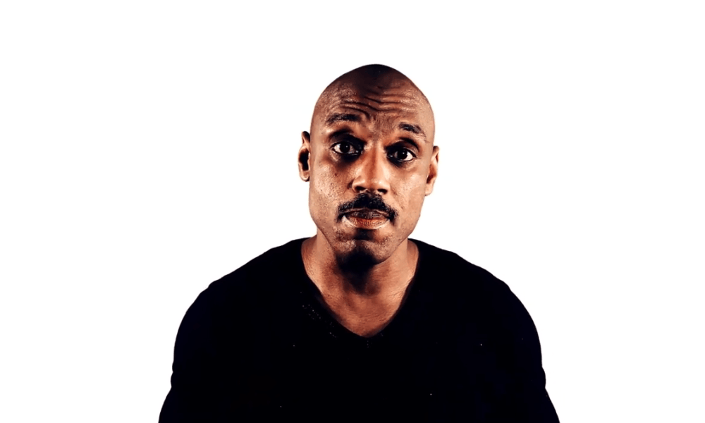 A person with a shaved head and a mustache is wearing a black football shirt. They are looking directly at the camera against a plain white background.