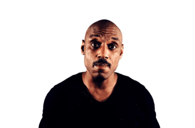 A person with a shaved head and a mustache is wearing a black football shirt. They are looking directly at the camera against a plain white background.