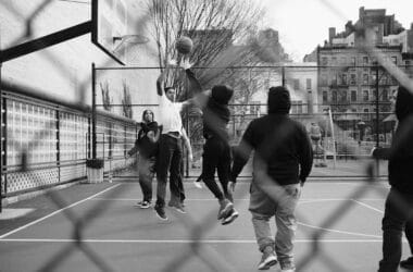 People enjoy a game of basketball on an outdoor court, viewed through a chain-link fence, channeling the spirit and precision of Reggie Miller.