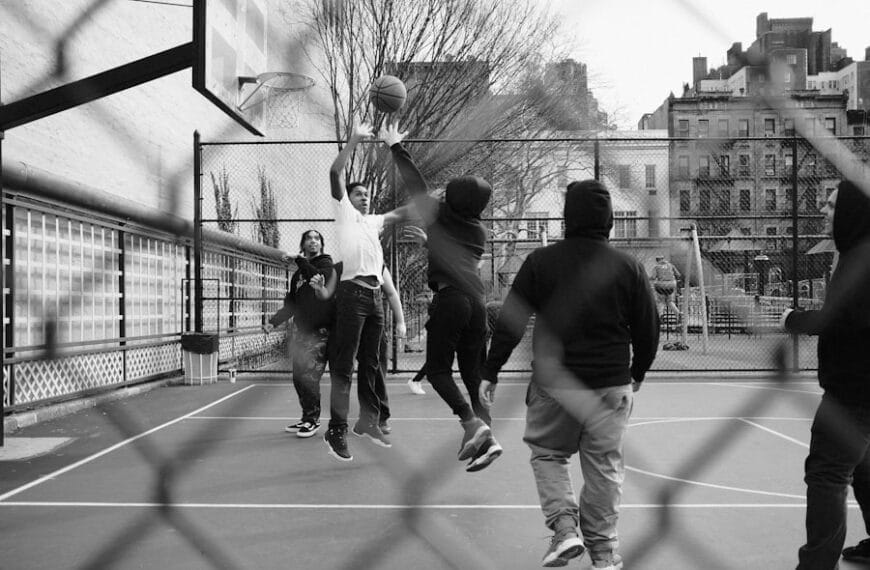 People enjoy a game of basketball on an outdoor court, viewed through a chain-link fence, channeling the spirit and precision of Reggie Miller.