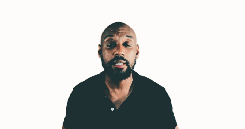A person with a beard in a black shirt, exuding a vibe more like football, stands confidently against a plain white background.