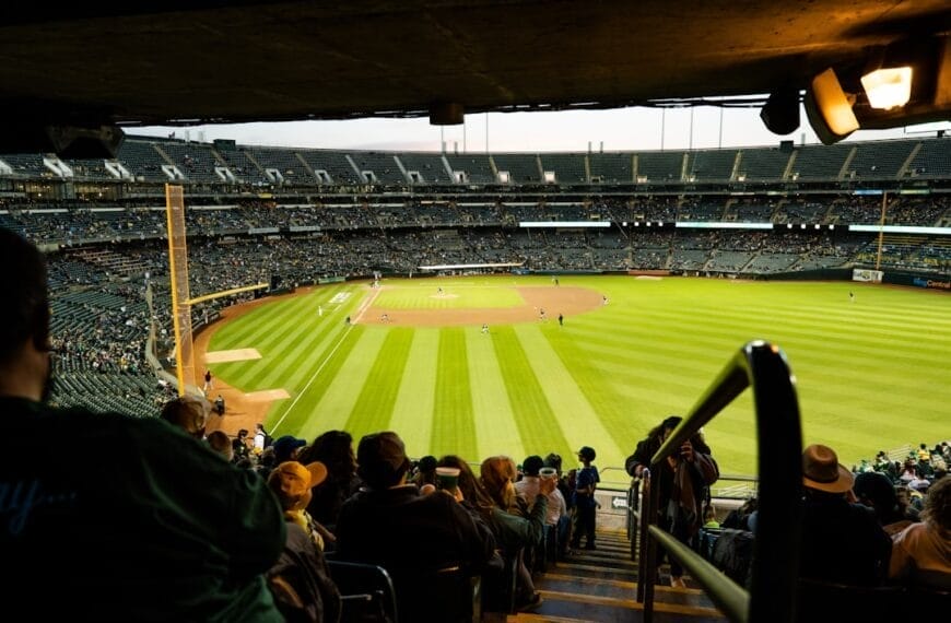 View from under a stadium overhang looking out onto a baseball field. The field is green with a dirt infield. Fans fill the stands, watching the game unfold against an overcast sky. With recent MLB rule changes enhancing the pace, excitement buzzes both on and off the field.