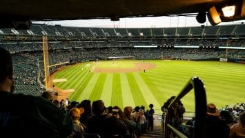 View from under a stadium overhang looking out onto a baseball field. The field is green with a dirt infield. Fans fill the stands, watching the game unfold against an overcast sky. With recent MLB rule changes enhancing the pace, excitement buzzes both on and off the field.
