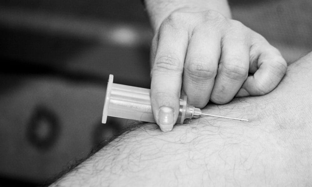 A hand injects a needle into an arm, administering a shot, reminiscent of the precision needed in forecasting NFL injuries. Black and white image.