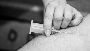 A hand injects a needle into an arm, administering a shot, reminiscent of the precision needed in forecasting NFL injuries. Black and white image.