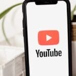 A smartphone, symbolizing digital influence, displays the YouTube logo on its screen. It stands upright, leaning against a white textured surface. A plant in a white pot casually peeks into the background.