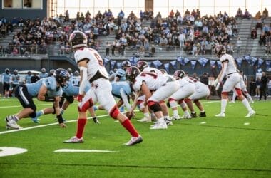 Two football teams face off on the field, with players in blue and white uniforms, while sports superfans fill the stands, creating an electrifying atmosphere.