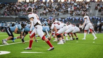 Two football teams face off on the field, with players in blue and white uniforms, while sports superfans fill the stands, creating an electrifying atmosphere.