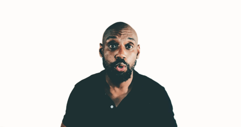 A surprised man in a black shirt stands against a plain white background, looking more like a football fan who's just witnessed an unbelievable goal.