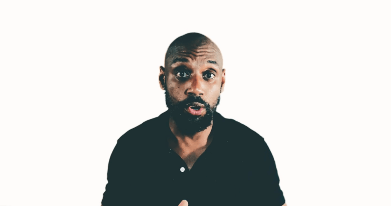 A surprised man in a black shirt gestures, as if celebrating a moment more like football, against a plain white background.