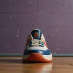 A single sneaker is placed on a wooden floor against a purple wall, echoing the style of 90's basketball. The shoe features a white body with blue and orange accents. A logo is visible on the tongue, while the soft lighting casts shadows that add to its nostalgic appeal.