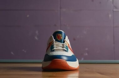 A single sneaker is placed on a wooden floor against a purple wall, echoing the style of 90's basketball. The shoe features a white body with blue and orange accents. A logo is visible on the tongue, while the soft lighting casts shadows that add to its nostalgic appeal.