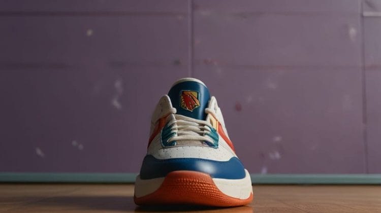 A single sneaker is placed on a wooden floor against a purple wall, echoing the style of 90's basketball. The shoe features a white body with blue and orange accents. A logo is visible on the tongue, while the soft lighting casts shadows that add to its nostalgic appeal.