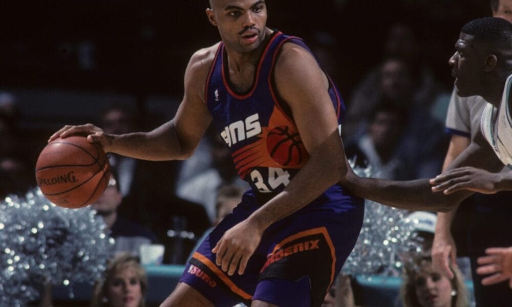 A basketball player in a purple jersey, reminiscent of the legendary Charles Barkley, holds the ball during an intense NBA game, with cheerleaders and a lively crowd in the background.