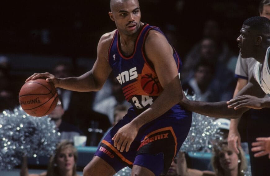 A basketball player in a purple jersey, reminiscent of the legendary Charles Barkley, holds the ball during an intense NBA game, with cheerleaders and a lively crowd in the background.