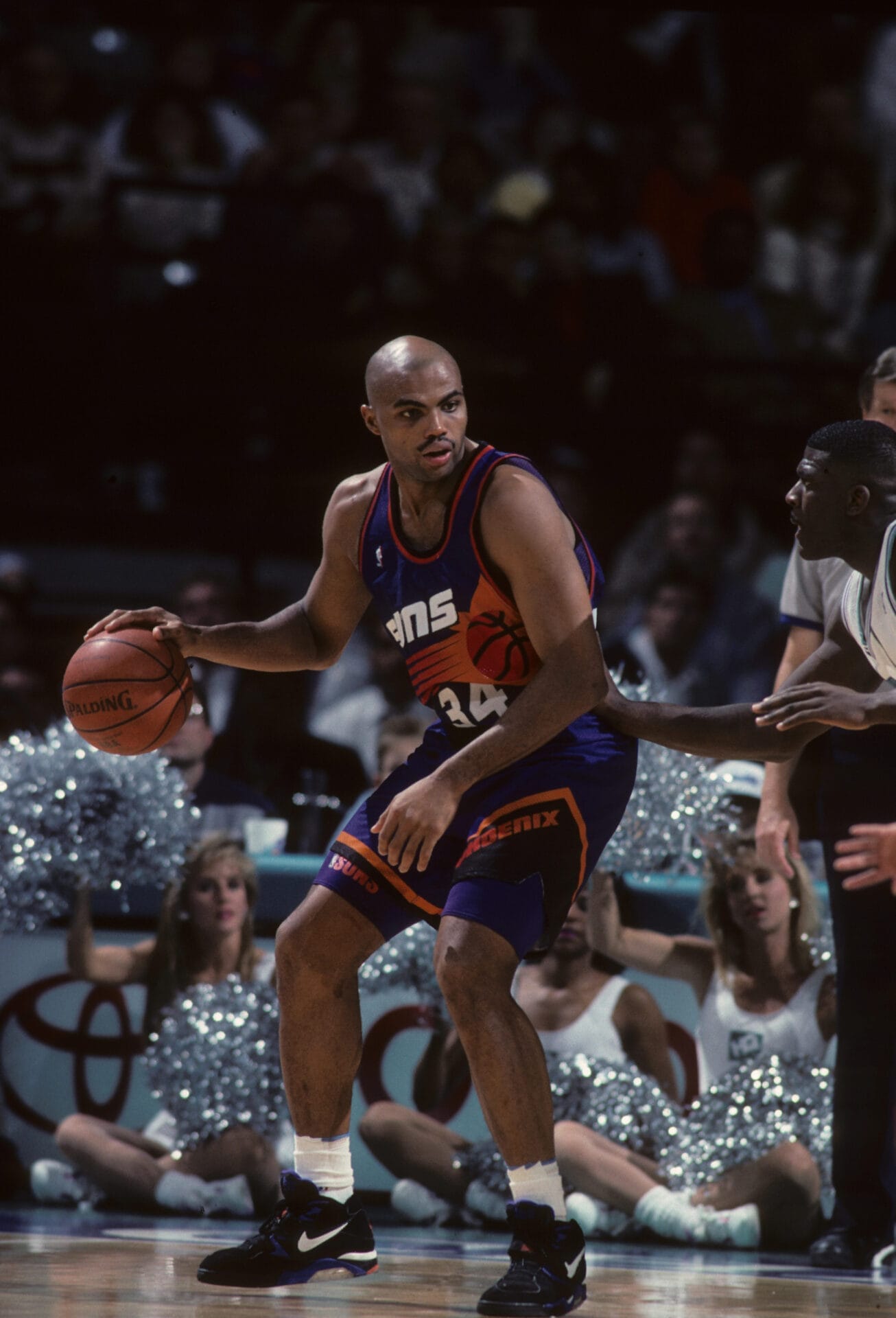 A basketball player in a purple jersey, reminiscent of the legendary Charles Barkley, holds the ball during an intense NBA game, with cheerleaders and a lively crowd in the background.