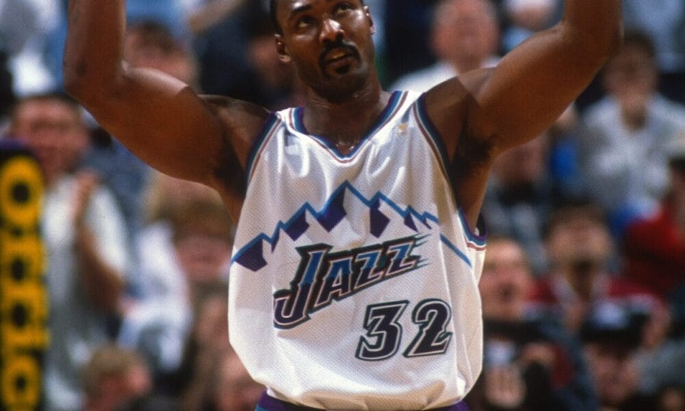 A basketball player in a white and purple jersey with the number 32 raises his arms during an NBA game, echoing the style of Karl Malone. The crowd buzzes with excitement in the background.