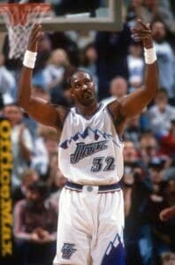 A basketball player in a white and purple jersey with the number 32 raises his arms during an NBA game, echoing the style of Karl Malone. The crowd buzzes with excitement in the background.