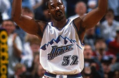 A basketball player in a white and purple jersey with the number 32 raises his arms during an NBA game, echoing the style of Karl Malone. The crowd buzzes with excitement in the background.