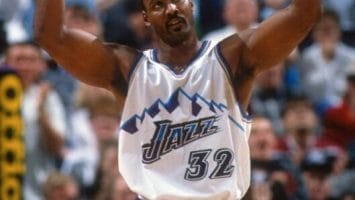 A basketball player in a white and purple jersey with the number 32 raises his arms during an NBA game, echoing the style of Karl Malone. The crowd buzzes with excitement in the background.